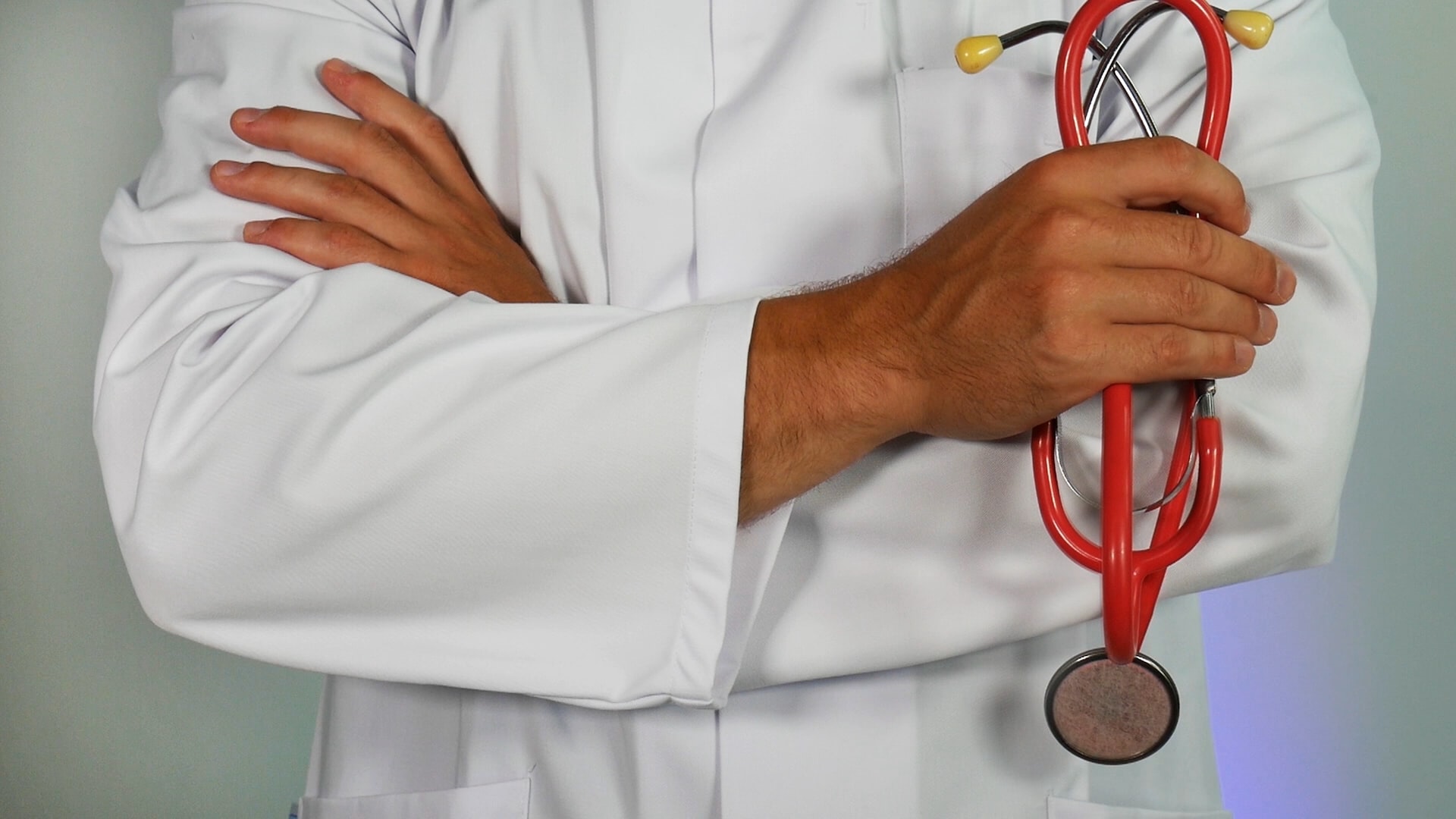 Un medico in camice bianco tiene in mano uno stetoscopio rosso e ha l’altro braccio incrociato.